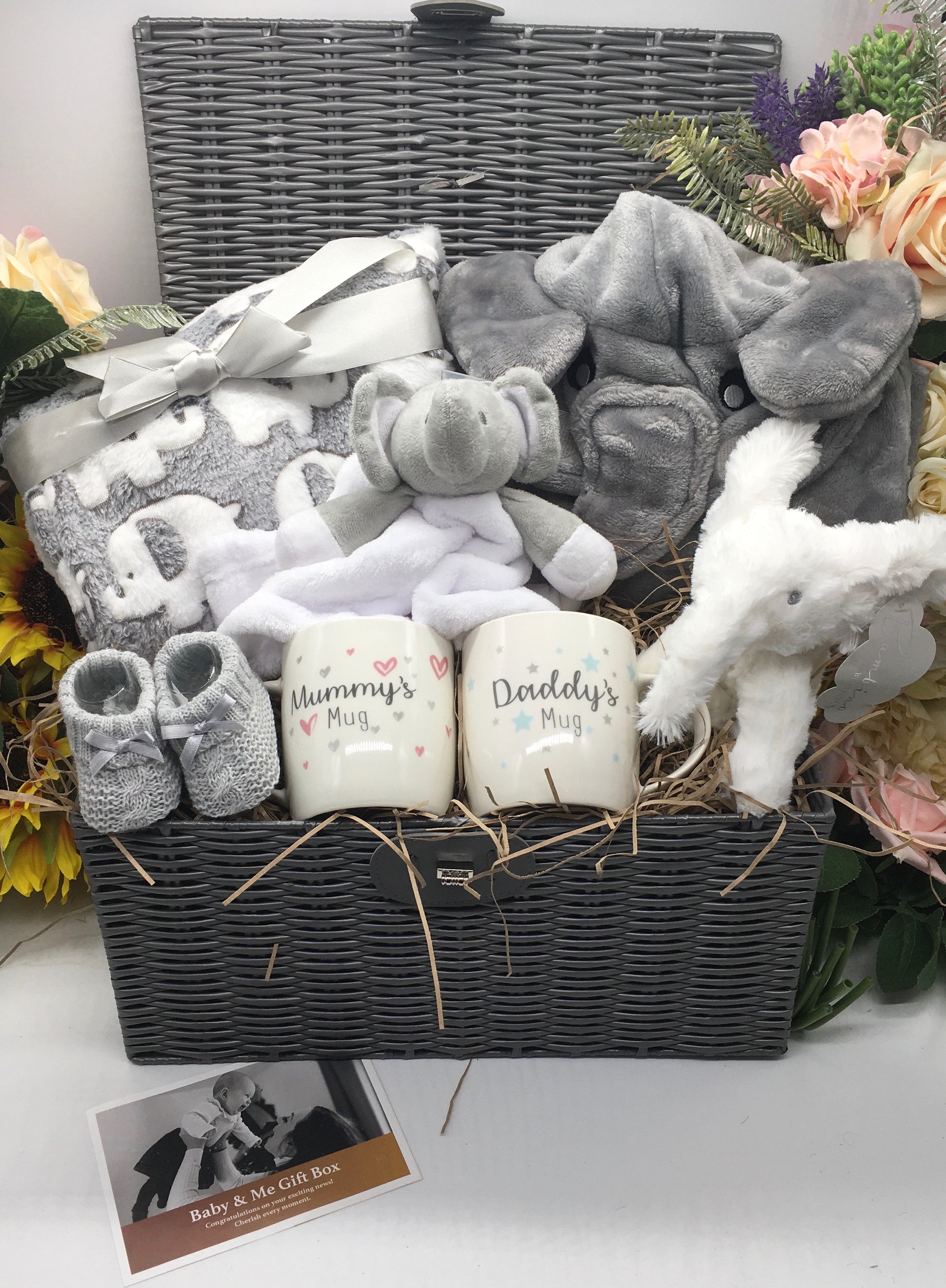 A deluxe baby hamper featuring a gray elephant dressing gown, a white and gray elephant baby wrap, a pair of white booties, a gray elephant comforter, and two mugs with 'Mummy's Mug' and 'Daddy's Mug' inscribed.